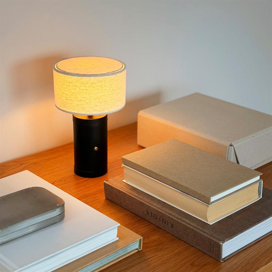 Bordlampe lyser blødt fra en rund, lys skærm på en mørk cylinderfod; står på et træbord omgivet af stakke bøger og pappkasser. Tekst på bogomslag: "KINFOLK".