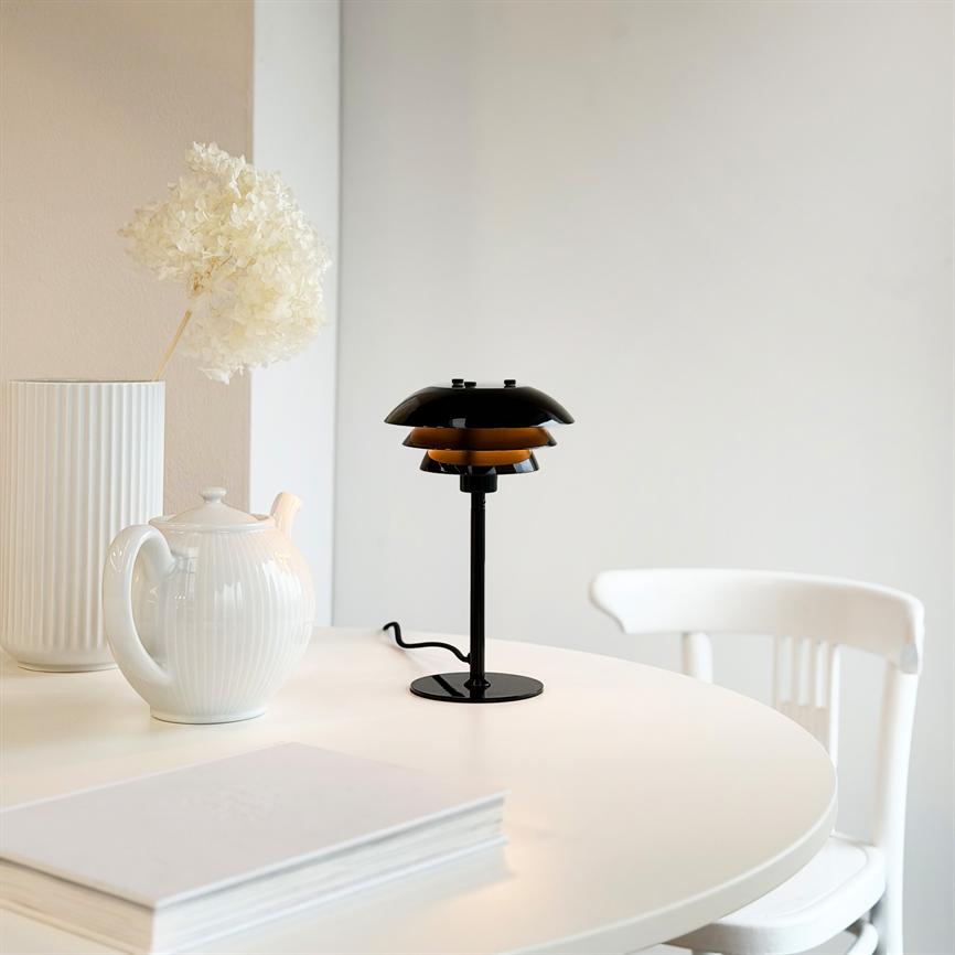 Den sorte bordlampe kaster et varmt skær og står på et rundt hvidt bord med en hvid tekande, en vase med tørret blomst og en hvid stol i et lyst rum.