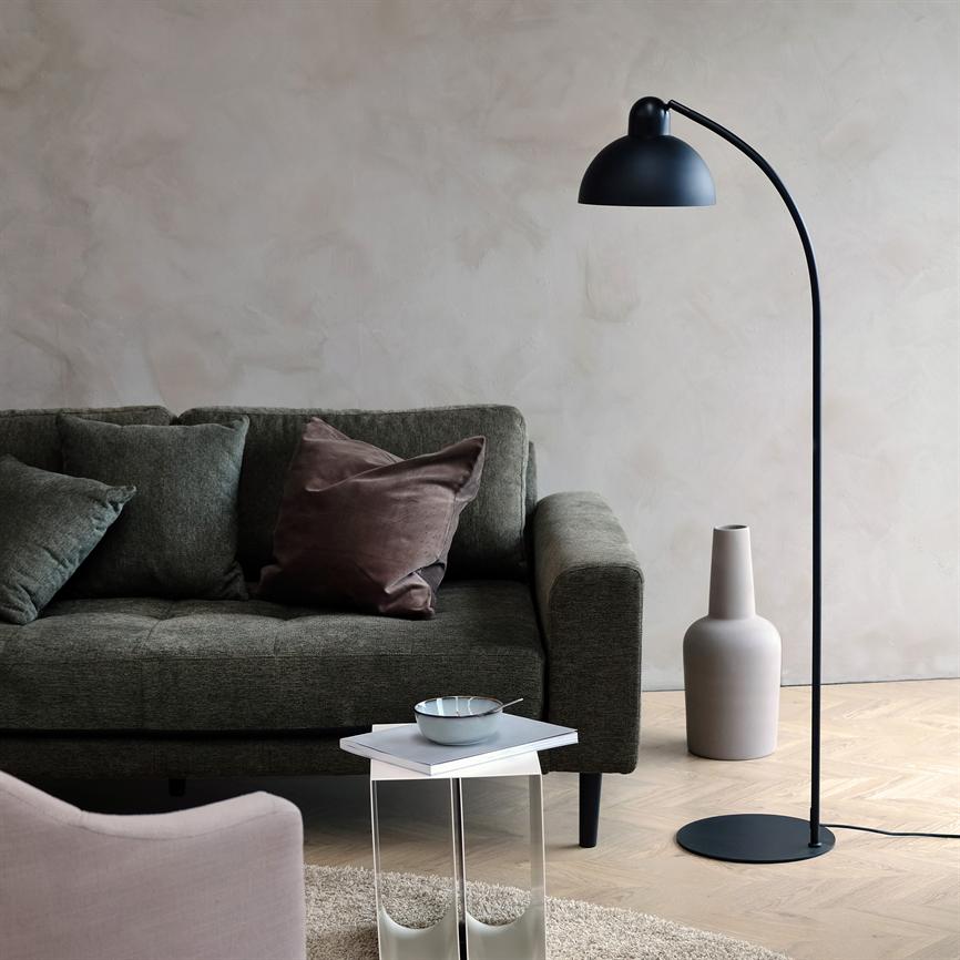 En sort, buet gulvlampe står ved siden af en mørkegrøn sofa med puder; et lille sidebord med skål står foran, i et lyst rum med pudret væg og trægulv.