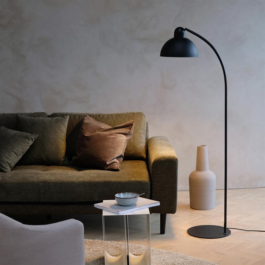 Sort bueformet gulvlampe kaster blødt lys over en olivengrøn sofa med puder, et lille sidebord med skål og en keramisk vase på lyst trægulv.